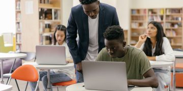 Students learning in a classroom setting with a teacher assisting and laptops on desks, creating an interactive education environment.