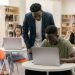 Students learning in a classroom setting with a teacher assisting and laptops on desks, creating an interactive education environment.