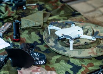 Compact white drone on camouflage gear, surrounded by flashlight and tools.
