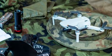 Compact white drone on camouflage gear, surrounded by flashlight and tools.
