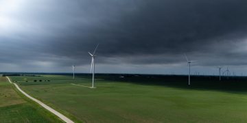 A stormy sky looms over a vast green landscape with wind turbines generating renewable energy.
