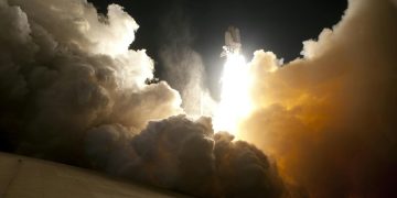 A dramatic image of a space shuttle launching into the night sky, surrounded by billowing smoke and intense light.