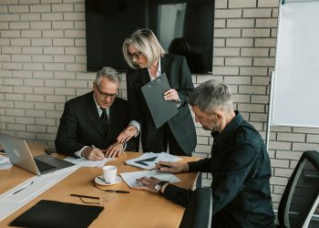 Three mature professionals in a business meeting discussing and signing documents in an office setting.
