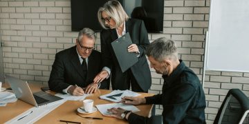 Three mature professionals in a business meeting discussing and signing documents in an office setting.