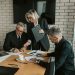 Three mature professionals in a business meeting discussing and signing documents in an office setting.