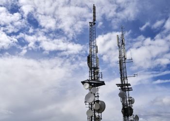Telecommunication towers with antennas and dishes under a bright cloudy sky.