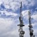 Telecommunication towers with antennas and dishes under a bright cloudy sky.