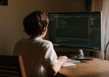 A teenager focused on coding software on a desktop monitor in a home office setting.