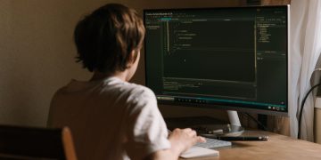 A teenager focused on coding software on a desktop monitor in a home office setting.