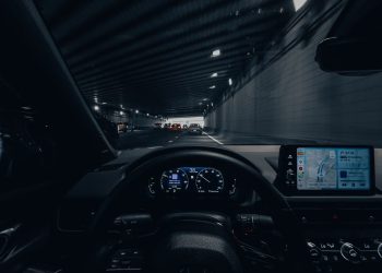 View from car dashboard driving through an illuminated city tunnel at night.