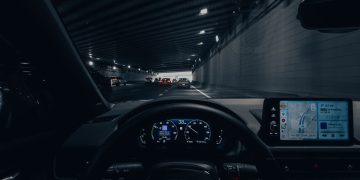 View from car dashboard driving through an illuminated city tunnel at night.