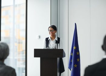 A woman giving a speech at a podium with an EU flag in a conference setting.