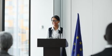 A woman giving a speech at a podium with an EU flag in a conference setting.
