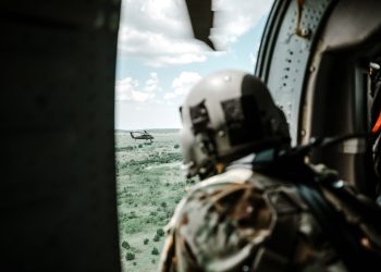 Helmeted soldier in helicopter observing another helicopter over a vast landscape.