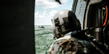 Helmeted soldier in helicopter observing another helicopter over a vast landscape.