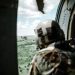 Helmeted soldier in helicopter observing another helicopter over a vast landscape.