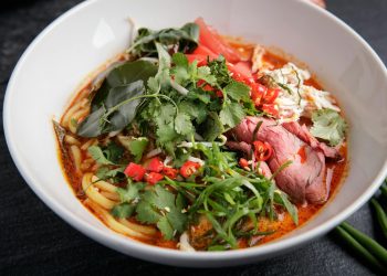 A vibrant and spicy Asian noodle soup featuring fresh herbs, beef, and chili peppers in a white bowl.