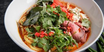 A vibrant and spicy Asian noodle soup featuring fresh herbs, beef, and chili peppers in a white bowl.