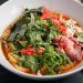 A vibrant and spicy Asian noodle soup featuring fresh herbs, beef, and chili peppers in a white bowl.