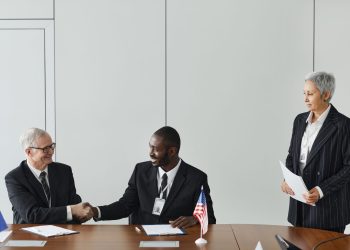 Multinational business meeting with agreement signing, featuring diverse professionals and flags.