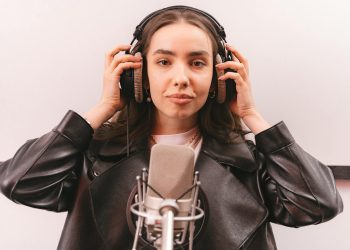 Woman in a leather jacket and headphones singing into a microphone in a studio.