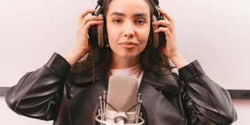 Woman in a leather jacket and headphones singing into a microphone in a studio.