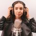 Woman in a leather jacket and headphones singing into a microphone in a studio.