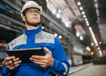 Factory worker in safety gear using a tablet in a well-lit industrial setting.