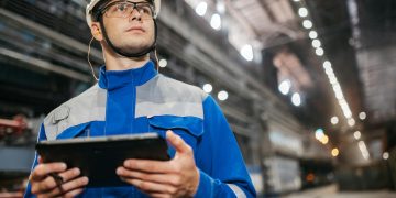 Factory worker in safety gear using a tablet in a well-lit industrial setting.