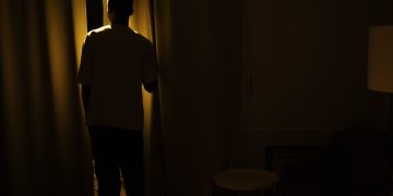 Silhouette of a person standing by a window in a dimly lit room, suggesting themes of solitude and contemplation.