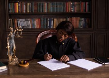 A judge in robes writing on a document at a desk in an office library with law books.