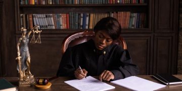 A judge in robes writing on a document at a desk in an office library with law books.