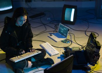 Young Asian woman engaged in computer hacking in a dimly lit, technologically equipped room.