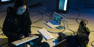 Young Asian woman engaged in computer hacking in a dimly lit, technologically equipped room.