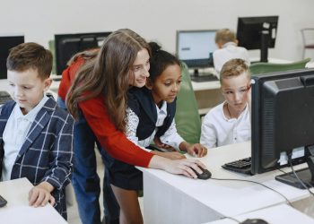 Teacher engaging with diverse children in a technology lesson using computers in a modern classroom.