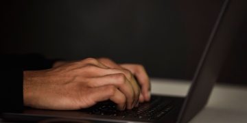 Crop unrecognizable person surfing internet on contemporary netbook and typing on keyboard in dark workspace