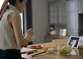 Asian woman preparing a fresh salad with tomatoes and lettuce in a contemporary kitchen setting.