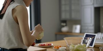 Asian woman preparing a fresh salad with tomatoes and lettuce in a contemporary kitchen setting.