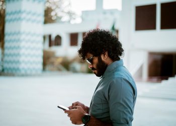 Side view of a curly-haired man with sunglasses using a smartphone in a sunny outdoor setting.