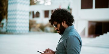 Side view of a curly-haired man with sunglasses using a smartphone in a sunny outdoor setting.