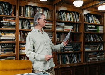 Elderly man reading a document in a cozy library setting surrounded by bookshelves.