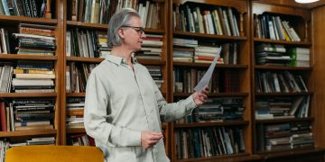 Elderly man reading a document in a cozy library setting surrounded by bookshelves.