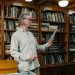 Elderly man reading a document in a cozy library setting surrounded by bookshelves.