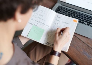An adult writing in a notebook with a laptop on a wooden table for work or leisure.