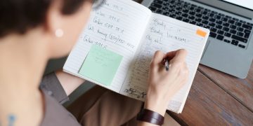 An adult writing in a notebook with a laptop on a wooden table for work or leisure.