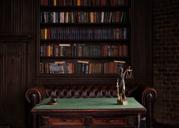 A classic study room with shelves of books, a gavel, and Lady Justice figurine on a green table.