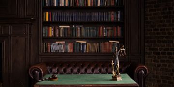 A classic study room with shelves of books, a gavel, and Lady Justice figurine on a green table.