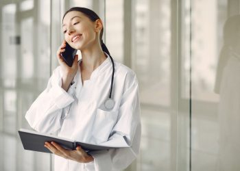 Smiling ethnic female doctor in medical uniform with stethoscope and notebook talking on smartphone while standing in gl