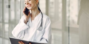 Smiling ethnic female doctor in medical uniform with stethoscope and notebook talking on smartphone while standing in gl