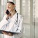 Smiling ethnic female doctor in medical uniform with stethoscope and notebook talking on smartphone while standing in gl